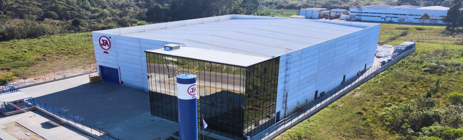 Fábrica da JEA Sul vista do alto: galpão branco com a entrada de vidro espelhado, o logo da JEA Sul na parede e a torre azul com o logo na frente.