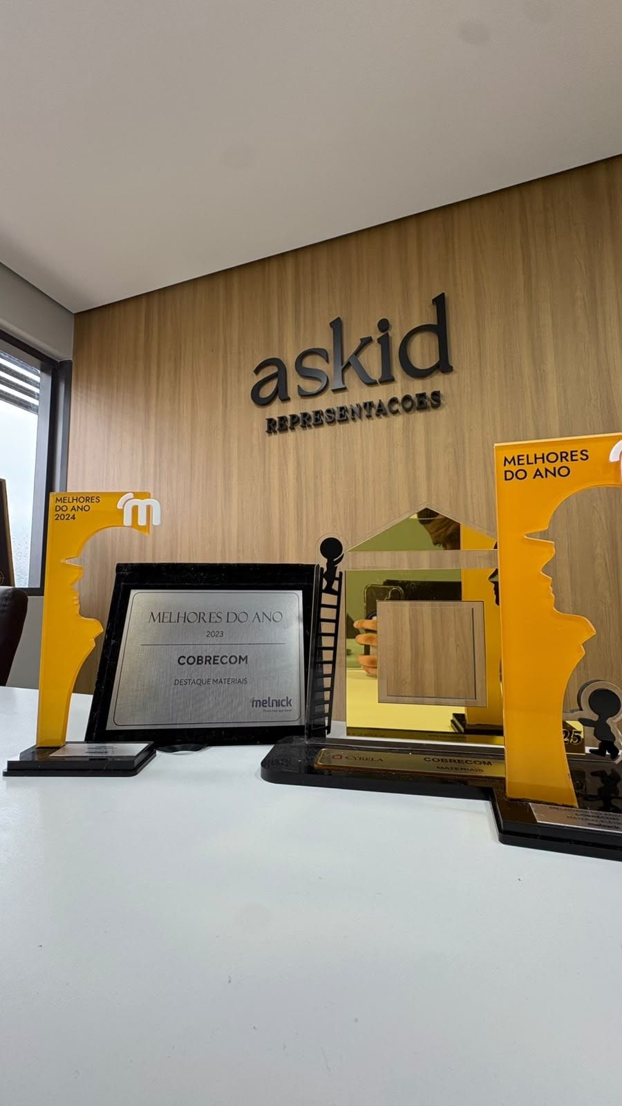 Troféus e placas de premiação sobre a mesa do escritório da ASKID, com a placa da empresa na parede ao fundo.